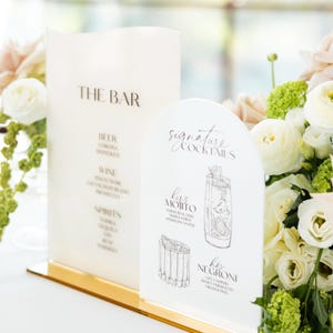 May include: Two white acrylic bar signs with gold bases. One sign reads "THE BAR" with a list of drinks. The other sign says "Signature Cocktails" and features illustrations of cocktails, including a Mojito and Negroni.