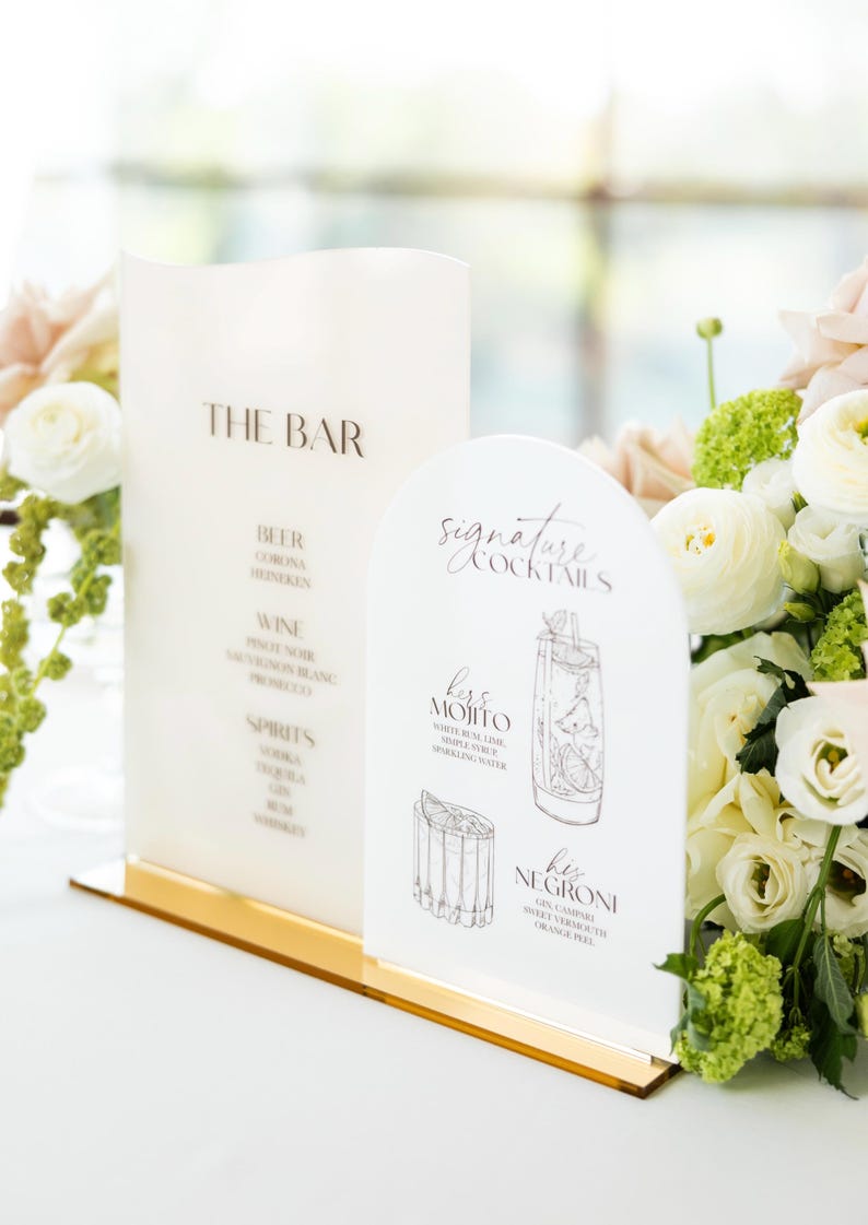 May include: Two white acrylic bar signs with gold bases. One sign reads "THE BAR" with a list of drinks. The other sign says "Signature Cocktails" and features illustrations of cocktails, including a Mojito and Negroni.