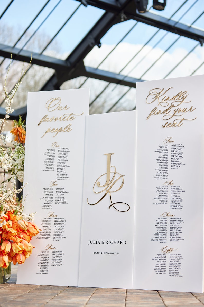 May include: White wedding seating chart with gold lettering. The chart is divided into sections labeled "One", "Two", "Three", "Four", and "Five". Each section lists names of guests. The chart also includes the couple's names, "Julia & Richard", and the date and location of the wedding, "05.31.24 | Newport, RI".