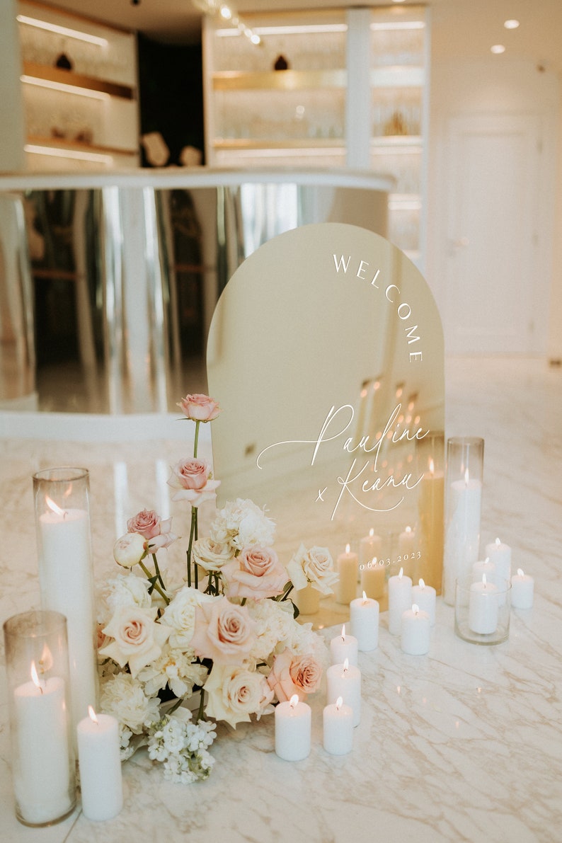 May include: A gold arch mirror with the words "Welcome Pauline & Keanu 08.03.2023" inscribed on it. The mirror is surrounded by white candles and a bouquet of white and pink roses.