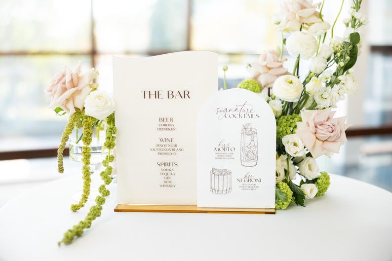 May include: A wedding bar menu with the text "THE BAR" and "signature cocktails" displayed on cream-colored cards. The menu lists beer, wine, and spirits. The cocktail menu features illustrations of drinks. Floral arrangements with pink and white roses surround the menu.