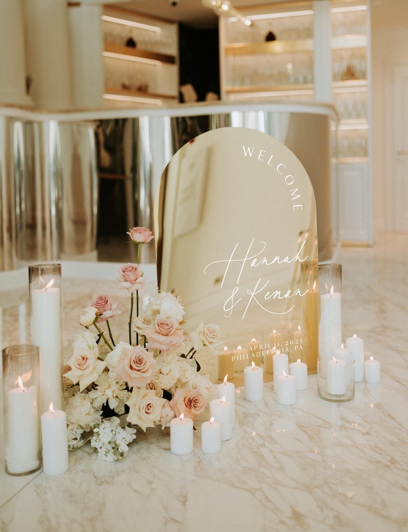 May include: A gold arch mirror with the words "Welcome Hannah & Kenan" and the date "April 1, 2023 Philadelphia, PA" written on it. The mirror is surrounded by white candles and a bouquet of pink and white flowers.