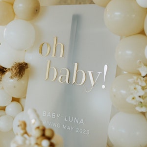 May include: A baby shower sign with the words "oh baby!" in gold lettering. The sign also includes the text "Baby Luna arriving May 2023". The sign is surrounded by white and beige balloons, with a decorated cake in the foreground.