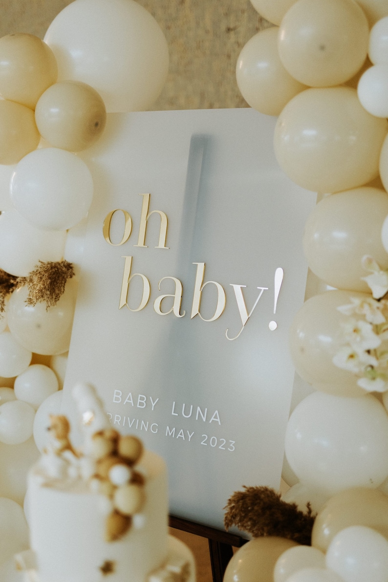May include: A baby shower sign with the words "oh baby!" in gold lettering. The sign also includes the text "Baby Luna arriving May 2023". The sign is surrounded by white and beige balloons, with a decorated cake in the foreground.