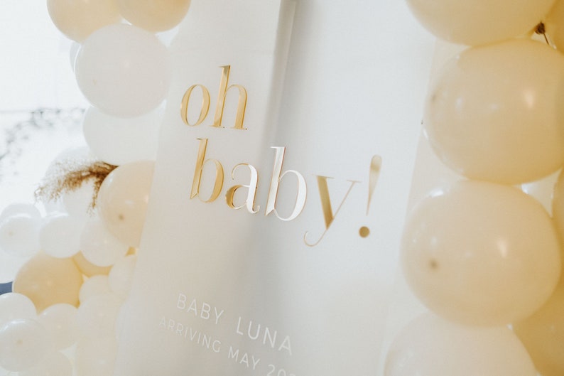 May include: A white sign with gold lettering that reads "oh baby!" is surrounded by white and cream-colored balloons. The sign also includes the text "BABY LUNA ARRIVING MAY 2023".