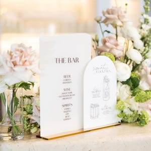 May include: A clear acrylic bar menu with gold trim, displaying "THE BAR" and drink options like beer, wine, and spirits. A separate sign lists signature cocktails. Floral arrangements of white and pink flowers frame the signs.