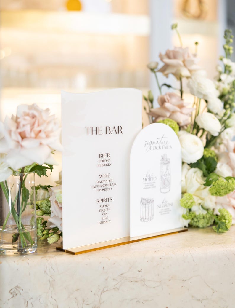 May include: A clear acrylic bar menu with gold trim, displaying "THE BAR" and drink options like beer, wine, and spirits. A separate sign lists signature cocktails. Floral arrangements of white and pink flowers frame the signs.