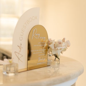 May include: A gold mirror arch sign with the text "Ring Ring" and instructions for leaving a message for the happy couple. The sign is on a white counter with a small vase of white roses.