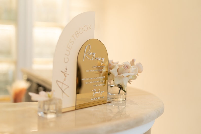 May include: A gold mirror arch sign with the text "Ring Ring" and instructions for leaving a message for the happy couple. The sign is on a white counter with a small vase of white roses.