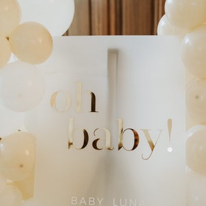 May include: A white sign with gold lettering that reads "oh baby!" is surrounded by white and cream-colored balloons. The sign also includes the name "BABY LUNA" at the bottom, perfect for a baby shower or gender reveal party.