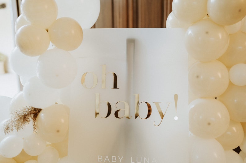 May include: A white sign with gold lettering that reads "oh baby!" is surrounded by white and cream-colored balloons. The sign also includes the name "BABY LUNA" at the bottom, perfect for a baby shower or gender reveal party.