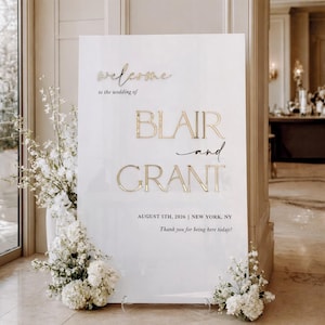 Op de afbeelding: Een wit welkomstbord voor een bruiloft met gouden letters met de tekst "welcome" en "Blair and Grant". Het bord bevat de datum 5 augustus 2026 en de locatie New York, NY. Witte bloemen zijn aan de voet van het bord geplaatst.
