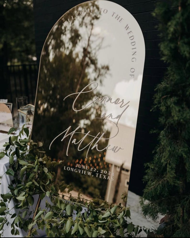 May include: A gold arch mirror with the text "Welcome to the Wedding of Conner and Matthew June 22, 2021 Longview, Texas" etched into the surface.