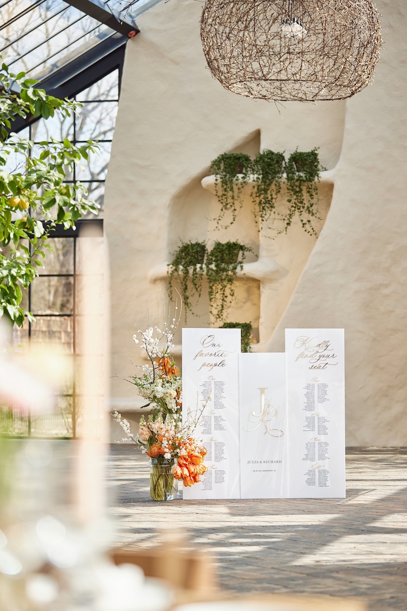 May include: White wedding seating chart with gold calligraphy. The chart is divided into three sections, with the left section titled "Our favorite people", the middle section titled "Julia & Richard", and the right section titled "Kindly find your seat".