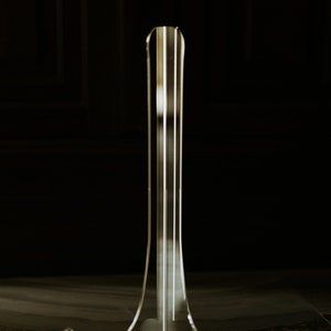 May include: A clear acrylic display stand with a tall, slender base and three curved arms. The stand is designed to hold and display items such as plates, photos, or artwork.