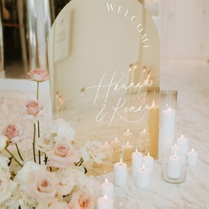 May include: A gold arch-shaped mirror with the words "Welcome Hannah & Kenan" and the date "April 11, 2025 Philadelphia, PA" written on it. The mirror is surrounded by white candles and a floral arrangement.