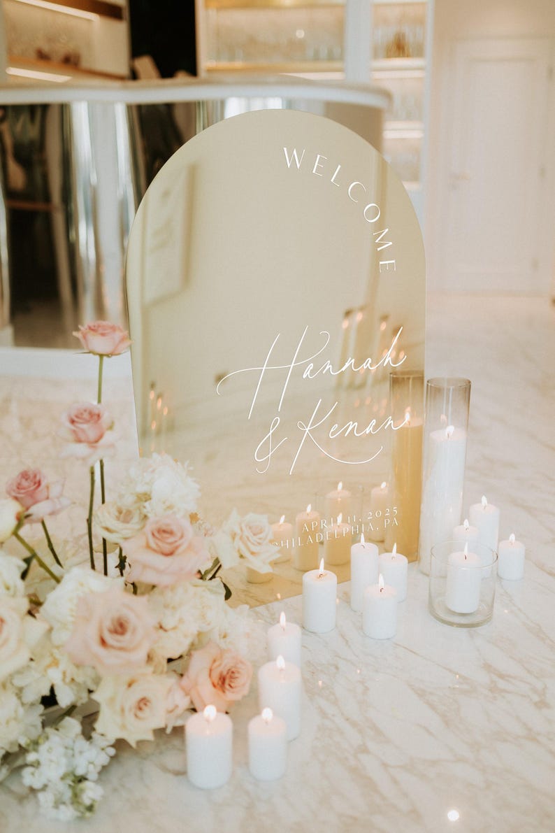 May include: A gold arch-shaped mirror with the words "Welcome Hannah & Kenan" and the date "April 11, 2025 Philadelphia, PA" written on it. The mirror is surrounded by white candles and a floral arrangement.