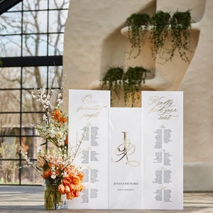 May include: Two white wedding seating charts with gold calligraphy. The left chart says "Our favorite people" and the right chart says "Kindly find your seat". A gold monogram of "J & R" is in the center. The charts are on a wooden floor with a vase of orange and white flowers.