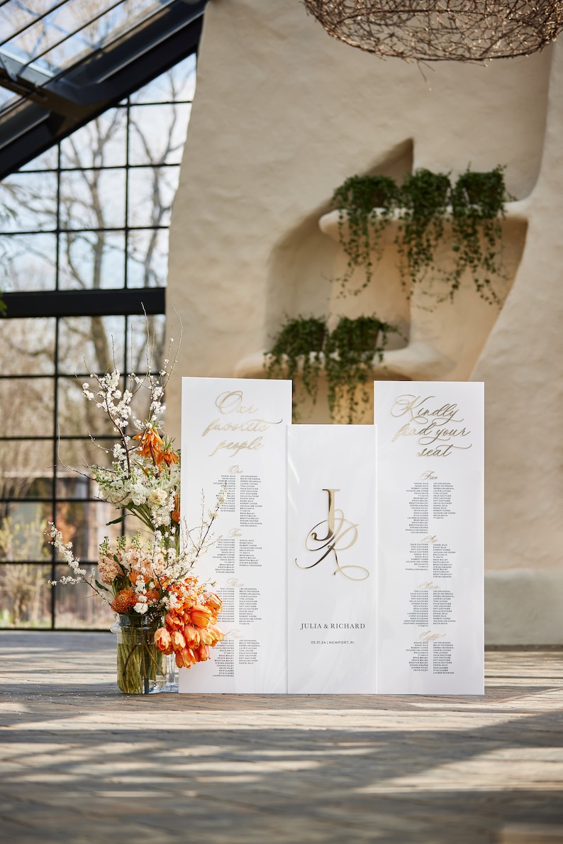 May include: Two white wedding seating charts with gold calligraphy. The left chart says "Our favorite people" and the right chart says "Kindly find your seat". A gold monogram of "J & R" is in the center. The charts are on a wooden floor with a vase of orange and white flowers.