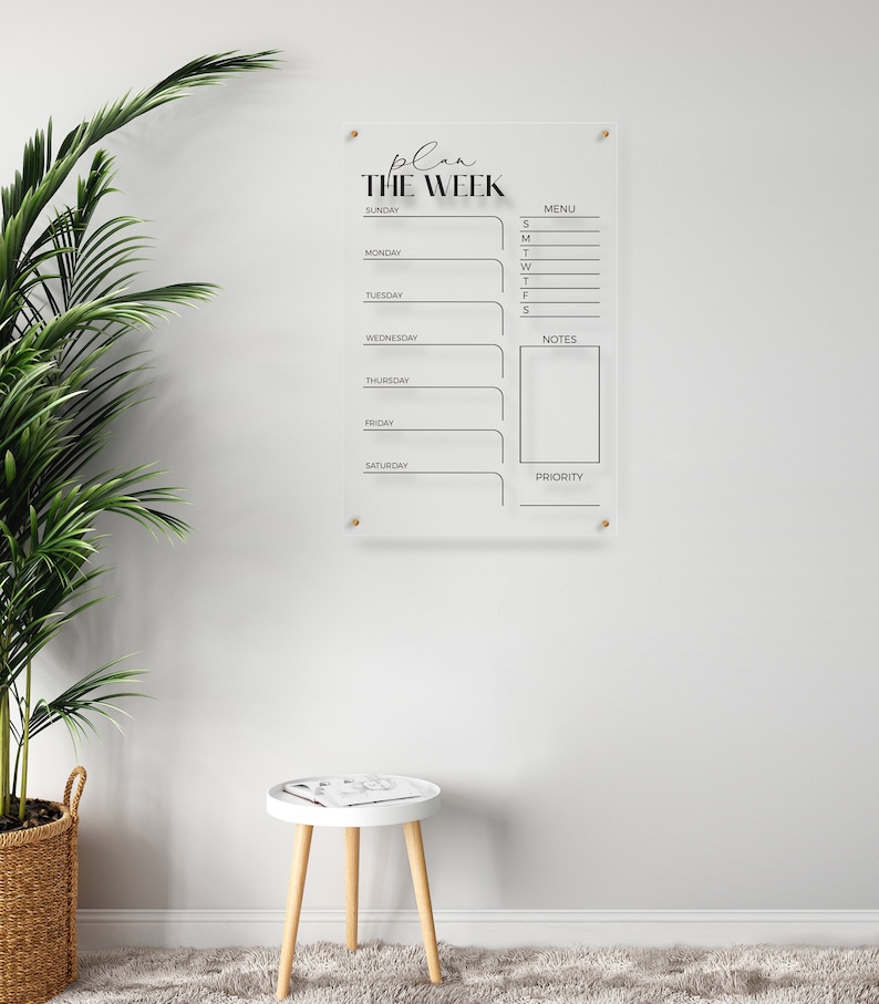 White wall with plant in basket on the left and a 3 legged chair on a shaggy carpet. On the wall in the centre there is a large acrylic panel with black lettering
