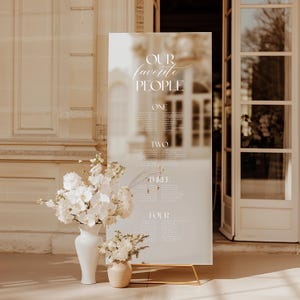 Puede incluir: Un plano de asientos de boda de acrílico esmerilado con el texto "OUR favorite PEOPLE" en una elegante escritura. El gráfico se muestra en un soporte dorado, con un fondo borroso de un edificio y puertas abiertas. Arreglos florales blancos en jarrones en primer plano.