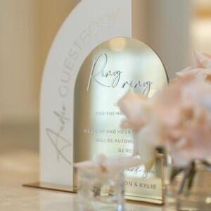 May include: A white acrylic arch with the words "Jade Guestbook" and "Ring Ring" inscribed on it. The arch is placed on a gold stand and has a mirror inside. The mirror has the text "For the happy couple, hang up and your ring will be automatically be returned. Thank you, [Name] & [Name]"