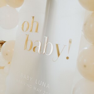 May include: A white sign with gold lettering that reads "oh baby!" is surrounded by white and cream-colored balloons. The sign also includes the text "BABY LUNA ARRIVING MAY 2023".