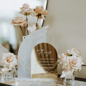 May include: A gold and white acrylic guest book sign with the text "Audio Guestbook" and "ring ring". The sign has instructions for guests to record a message for the couple. The sign is decorated with pink roses.