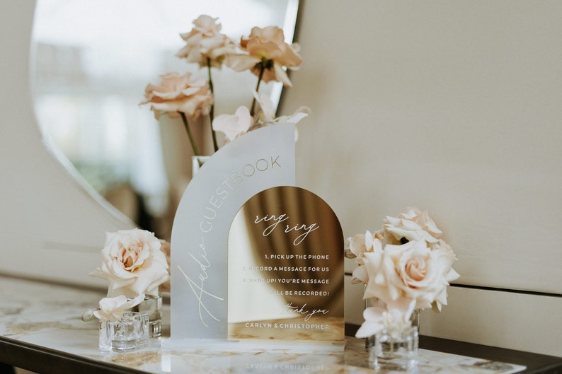 May include: A gold and white acrylic guest book sign with the text "Audio Guestbook" and "ring ring". The sign has instructions for guests to record a message for the couple. The sign is decorated with pink roses.