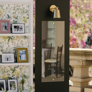 May include: A wedding seating chart with a black background and a gold sconce. The chart has names listed in white text. A white shelf displays framed photos and floral wallpaper. The initials "AJ" are at the top.