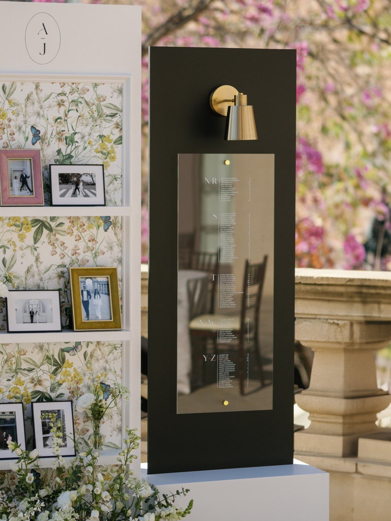 May include: A wedding seating chart with a black background and a gold sconce. The chart has names listed in white text. A white shelf displays framed photos and floral wallpaper. The initials "AJ" are at the top.
