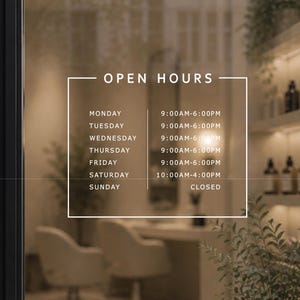 以下が含まれることがあります： 黒いフレームのガラスドアに、サロンの営業時間を示す「OPEN HOURS」の文字が白で表示されています。ガラス越しに見える内装は、ボトルや花瓶などの装飾品を備えた、ニュートラルな色調のミニマリストデザインです。