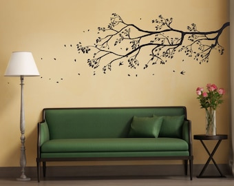 May include: A green sofa with two pillows sits in front of a wall with a black silhouette of a tree branch with leaves and birds. A lamp and a small table with a vase of pink flowers are also in the image.