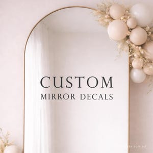 May include: An arched mirror with a gold frame. The mirror has the words "CUSTOM MIRROR DECALS" in black. The top right corner is decorated with dried flowers and cream-coloured orbs.