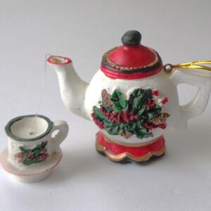 May include: A miniature ceramic teapot and teacup set. The teapot has a red lid and base, with a white body decorated with a green and red floral design. The teacup is white with a matching floral design. A gold string is attached to the teapot.