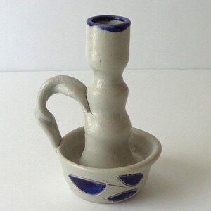 May include: A light gray ceramic candle holder with a handle and a bowl-shaped base. The rim of the candle holder and the base are decorated with hand-painted dark blue leaf-shaped accents. The piece has a smooth, matte finish throughout.