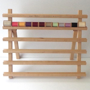 May include: A wooden thread rack with multiple shelves, holding spools of colorful thread. The rack is made of light-colored wood and has a simple, functional design. The spools of thread are in various colors, including pink, yellow, purple, and green.