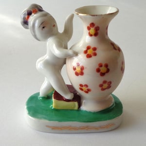 May include: A ceramic figurine of a cherubic figure with a vase. The vase is decorated with red and yellow flowers. The figure is white, and the base is green with gold trim. The figurine is on a white surface.