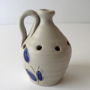 May include: A light gray ceramic jug with a single handle and a narrow neck. The body features a series of small circular holes around the upper section and a decorative pattern of three glossy blue leaf-shaped indentations on the lower side.