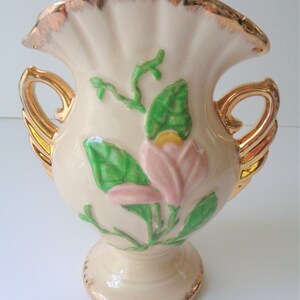 May include: A cream-colored ceramic vase with gold accents and a pink flower design. The vase has two gold handles and a ruffled top.