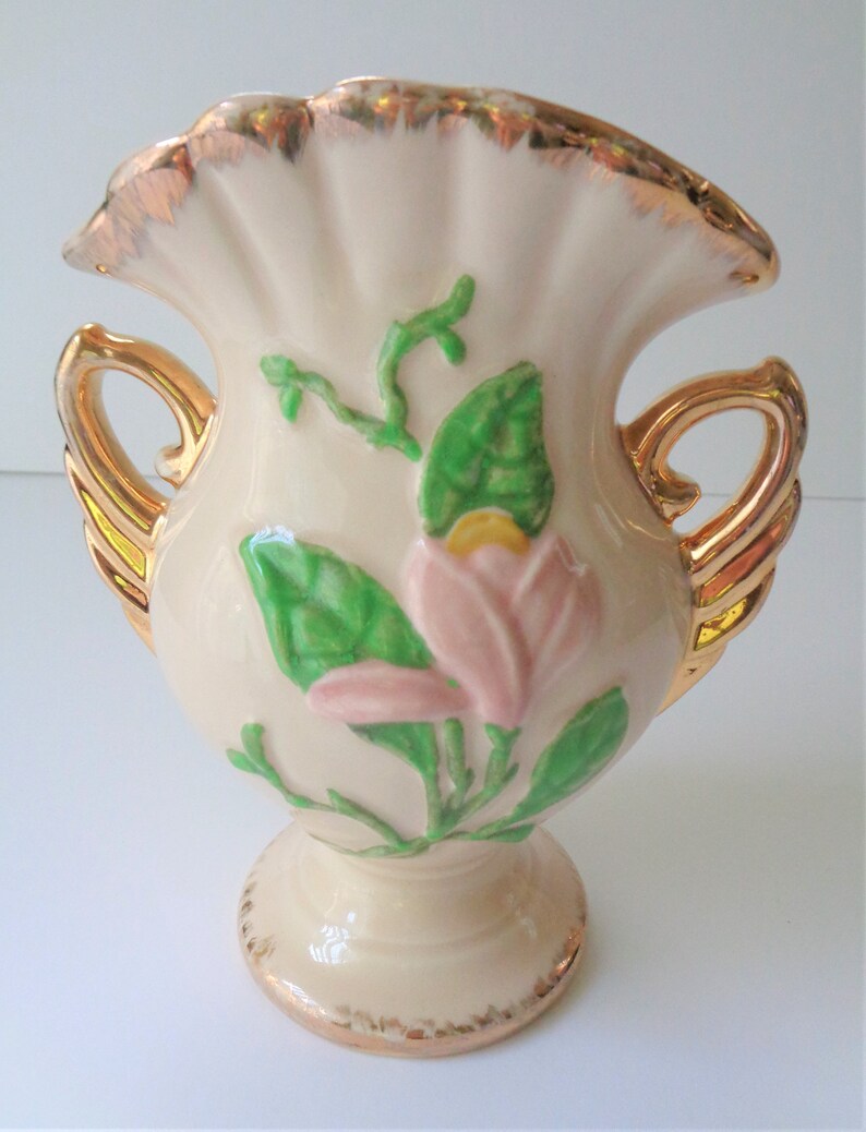 May include: A cream-colored ceramic vase with gold accents and a pink flower design. The vase has two gold handles and a ruffled top.