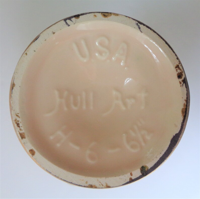 May include: The bottom of a ceramic vase with the words "USA Hull Art" and a number "K-618" in brown lettering on a light pink background.