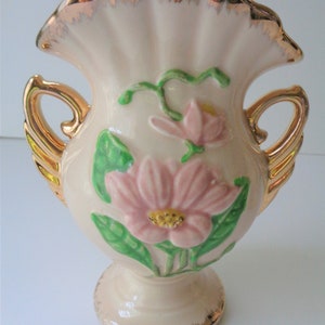 May include: A small, cream-colored ceramic vase with gold accents. The vase has two handles and features a floral design with pink and green flowers.