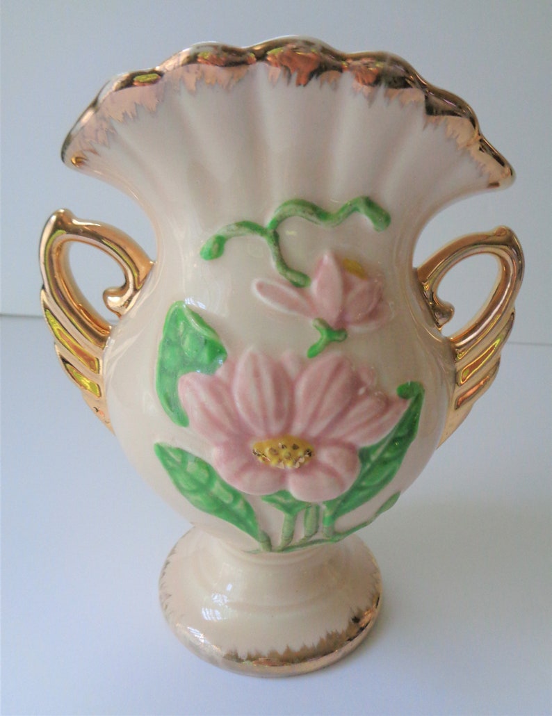 May include: A small, cream-colored ceramic vase with gold accents. The vase has two handles and features a floral design with pink and green flowers.