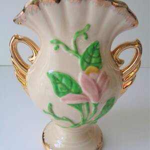 May include: A small, cream-colored ceramic vase with gold trim and handles. The vase features a pink and green floral design.
