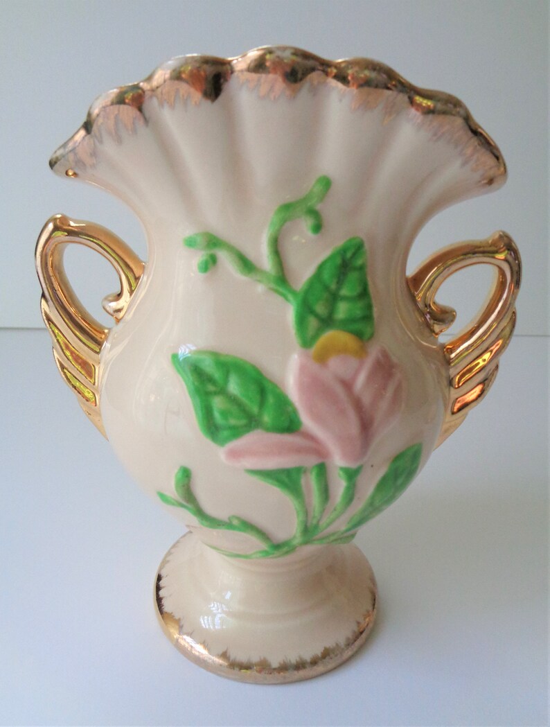 May include: A small, cream-colored ceramic vase with gold trim and handles. The vase features a pink and green floral design.