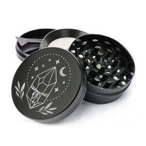 May include: Black metal grinder with a silver design of a crystal, moon, and stars. The grinder has a three-piece design with a screen and a herb catcher.