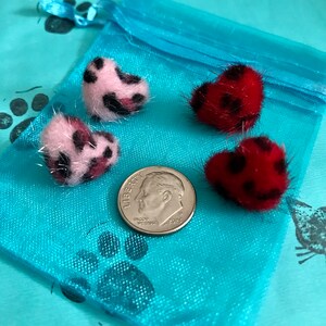 May include: Four heart-shaped, fuzzy ornaments with black spots, in pink and red, are displayed on a blue pouch next to a dime. The ornaments have a soft, textured appearance. The background is a light blue fabric with paw print designs.