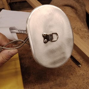 Op de afbeelding: Een zilverkleurige veldfles met een D-ringbevestiging. De veldfles is ovaal van vorm en lijkt van metaal te zijn. De achtergrond toont een houten oppervlak en andere objecten.