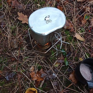 Op de afbeelding: Een metalen kampeerkooktoestel met een pot en deksel, buiten opgesteld. Het fornuis is aangestoken, met een kleine zwarte pan en een gele leren handschoen in de buurt. De scène speelt zich af op een bed van bladeren en twijgen.